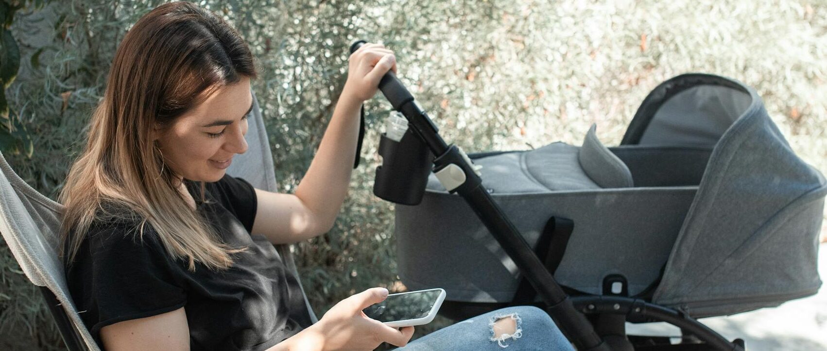 A young mother sits outdoors using a smartphone next to a stroller, enjoying a sunny day.
